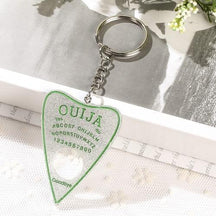 Silver glitter planchette keychain with green spirit-board lettering and a green outline, resting on white lace beneath a silver-tone chain and keyring.