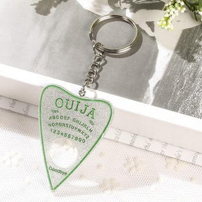 Silver glitter planchette keychain with green spirit-board lettering and a green outline, resting on white lace beneath a silver-tone chain and keyring.