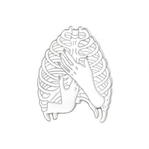 Silver-tone ribcage pin with an open skeletal outline and two hands crossed over the front.