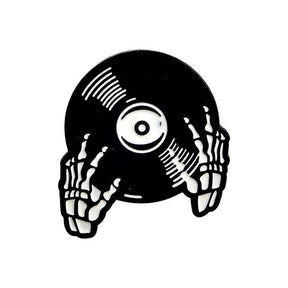 Black vinyl-record enamel pin held by illustrated white skeleton hands, with curved groove marks and a white circular centre label.