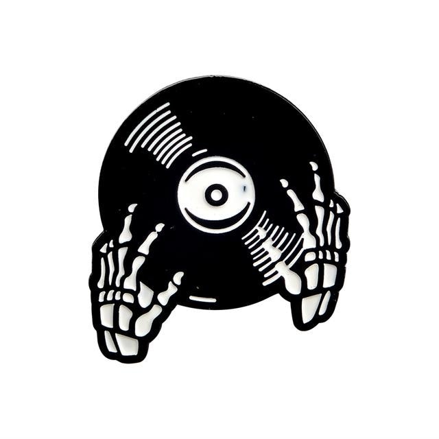 Black vinyl-record enamel pin held by illustrated white skeleton hands, with curved groove marks and a white circular centre label.