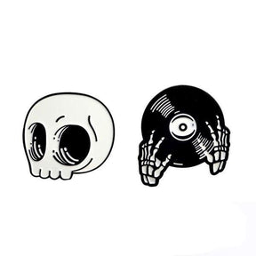 Two black-and-white enamel pin designs on white: a rounded skull with large eye sockets and skeleton hands holding a vinyl record.