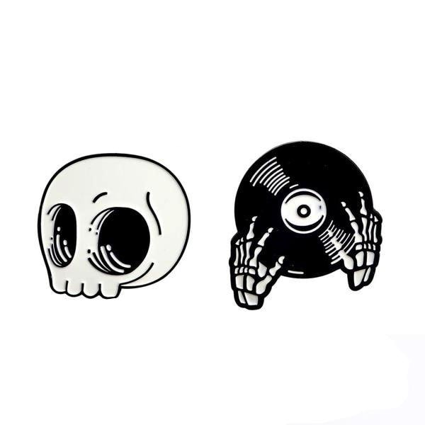 Two black-and-white enamel pin designs on white: a rounded skull with large eye sockets and skeleton hands holding a vinyl record.