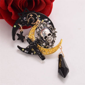 Close view of the layered skull cameo, black cross, small stars and gold-tone filigree moon, arranged beside a red rose. The pointed black charm hangs below the clip.