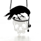 Black crow perched above a reflective skull pendant on a dark chain. A small black rose and faceted drop hang beside the skull.