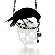 Black crow perched above a reflective skull pendant on a dark chain. A small black rose and faceted drop hang beside the skull.