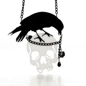 Black crow perched above a reflective skull pendant on a dark chain. A small black rose and faceted drop hang beside the skull.