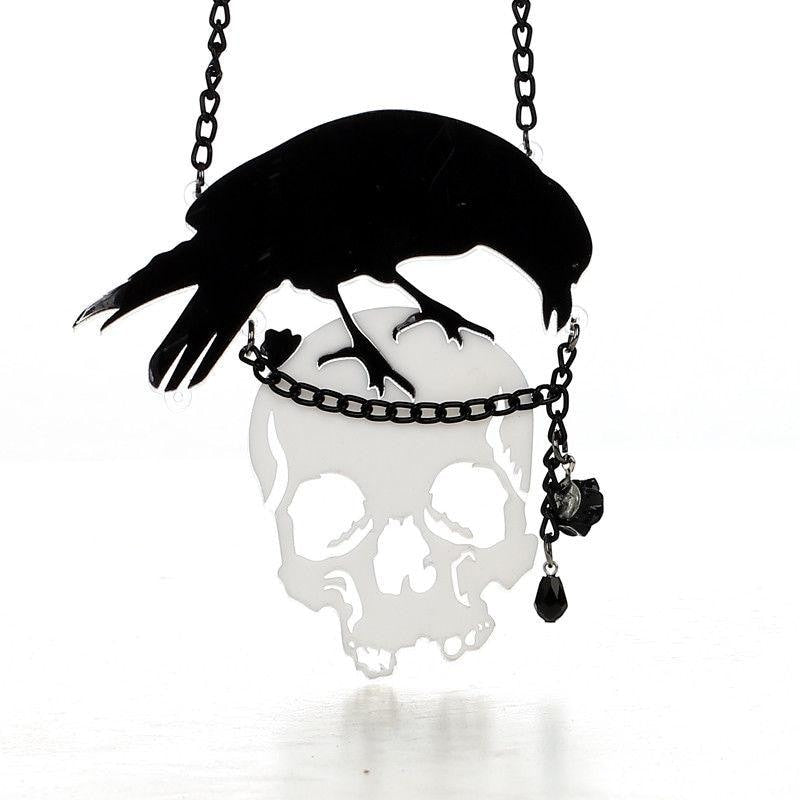 Black crow perched above a reflective skull pendant on a dark chain. A small black rose and faceted drop hang beside the skull.