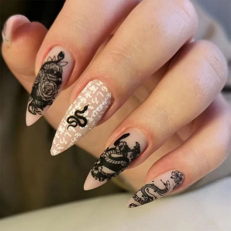 Pointed beige press-on nails with black snake and rose artwork, shown on a hand against a pale gray background. White lettering accents one nail.