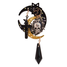 Black crescent moon hair clip on white, layered with gold-tone filigree, a silver-tone skull cameo, stars and a black cross. A faceted black charm hangs below.