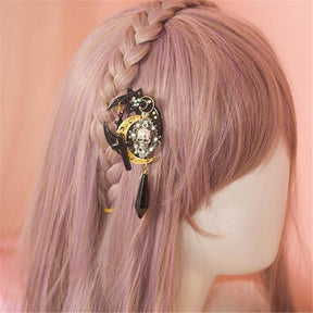 Crescent moon skull clip placed beside a braid in a pale pink wig. The gold-tone filigree and hanging black charm remain visible against the hair.