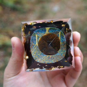 Tiger eye and obsidian orgone pyramid with a golden-brown sphere and blue Tree of Life inlay. Viewed from above on a palm, showing the tree disc beneath the round sphere.