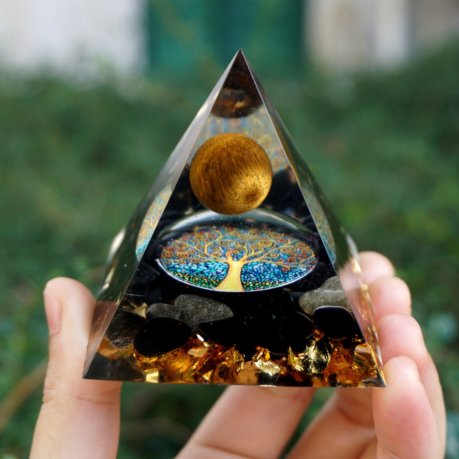 Additional view of Tiger eye and obsidian orgone pyramid with a golden-brown sphere and blue Tree of Life inlay. A full front view held farther from the camera shows the pointed outline and tree inlay.