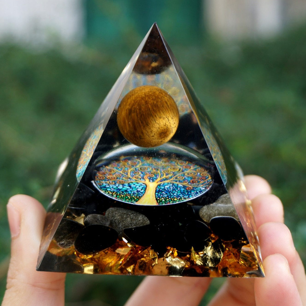 Tiger eye and obsidian orgone pyramid with a golden-brown sphere and blue Tree of Life inlay. Held upright outdoors with black stones and gold-tone flecks visible at the base.