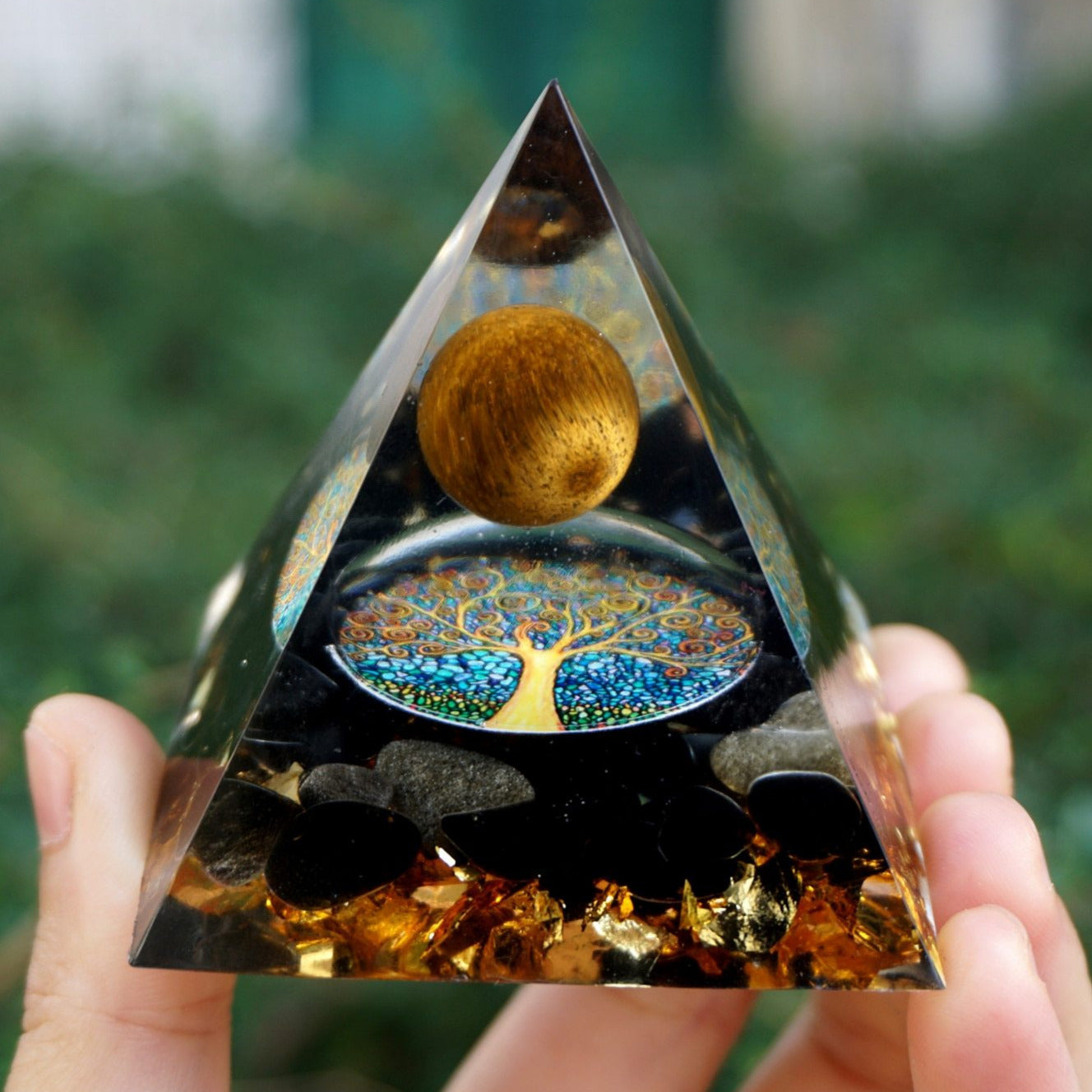 Tiger eye and obsidian orgone pyramid with a golden-brown sphere and blue Tree of Life inlay. Held upright outdoors with black stones and gold-tone flecks visible at the base.