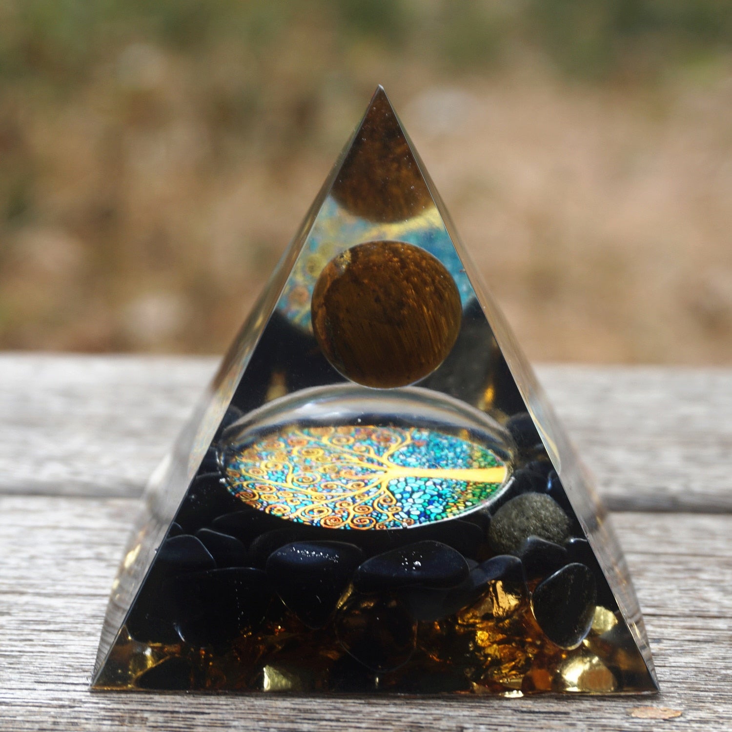 Tiger eye and obsidian orgone pyramid with a golden-brown sphere and blue Tree of Life inlay. Front view on wood with light reflecting from the clear tip and blue tree design.