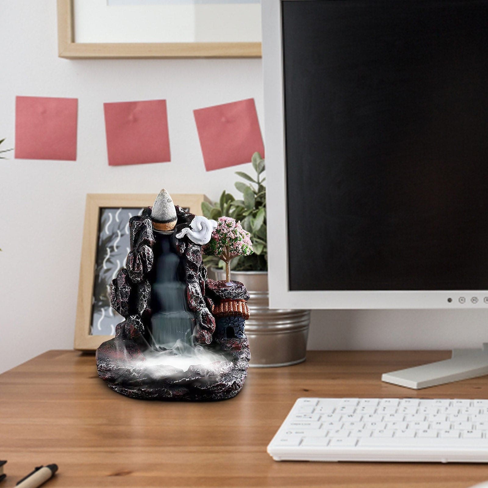 The miniature village burner displayed on a desk beside a keyboard and monitor, showing its house, pink tree and cascading smoke effect.