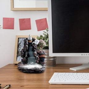 The miniature village burner displayed on a desk beside a keyboard and monitor, showing its house, pink tree and cascading smoke effect.