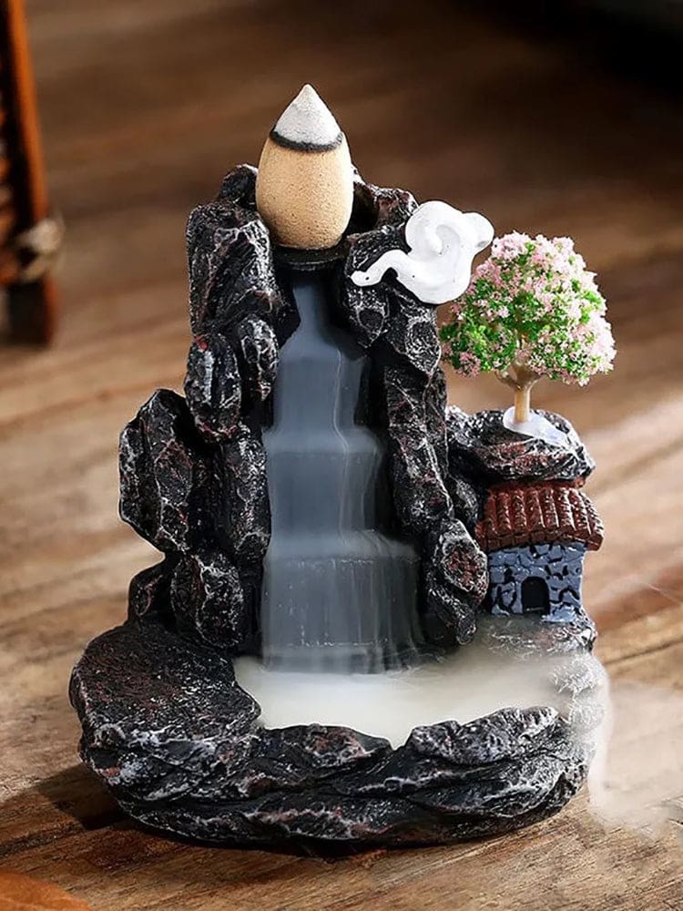 Mountain-village backflow incense burner on a wooden surface, with a tiled-roof house, pink blossom tree and white cloud beside a dark stepped smoke channel. Pale smoke descends toward the lower basin.