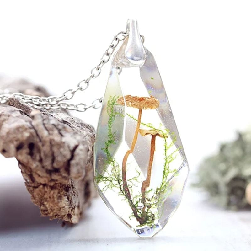 Clear faceted pendant suspended beside bark, showing warm brown toadstool caps on long thin stems and green botanical details inside.