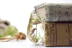 Wide still-life view of the toadstool necklace leaning against stacked books, with moss and mushroom props spread across the pale surface nearby.