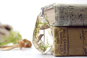 Toadstool pendant leaning against two old books, with the silver-tone chain crossing the top book and woodland props softly visible beside it.