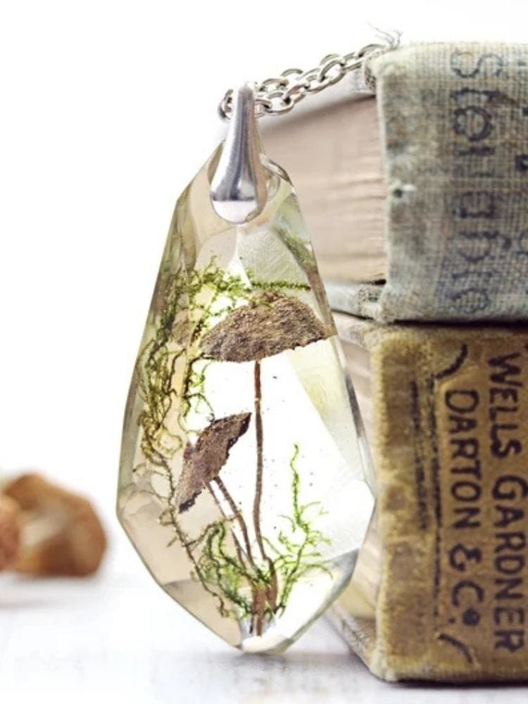 Clear faceted toadstool pendant standing beside stacked old books, with its silver-tone chain draped above the enclosed brown mushrooms and green strands.