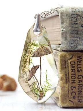 Front view of the transparent woodland pendant against a stack of clothbound books, showing mushroom caps, slender stems and green moss-like details.