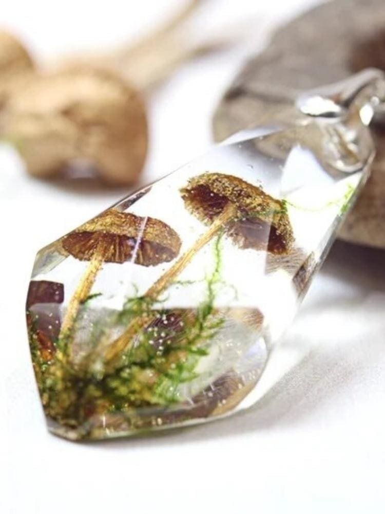 Close view of a clear faceted teardrop pendant holding tiny brown mushroom forms and fine green botanical details. The silver-tone bail and chain rest beside a wooden prop on a pale surface.