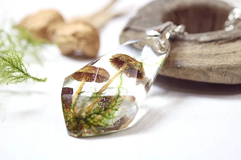 Angled close view of the faceted mushroom pendant lying beside a wooden dish, with the enclosed brown caps and green botanical strands visible through its clear body.