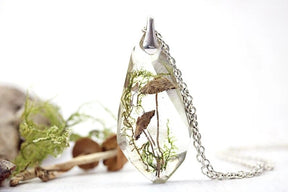Upright toadstool pendant with its silver-tone chain pooled beside it, photographed with moss and small mushroom props against white.