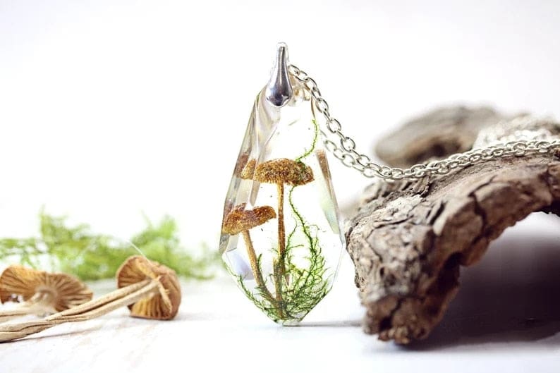 Clear mushroom terrarium pendant standing beside a piece of bark, with tiny mushrooms and green strands visible through the angular sides.