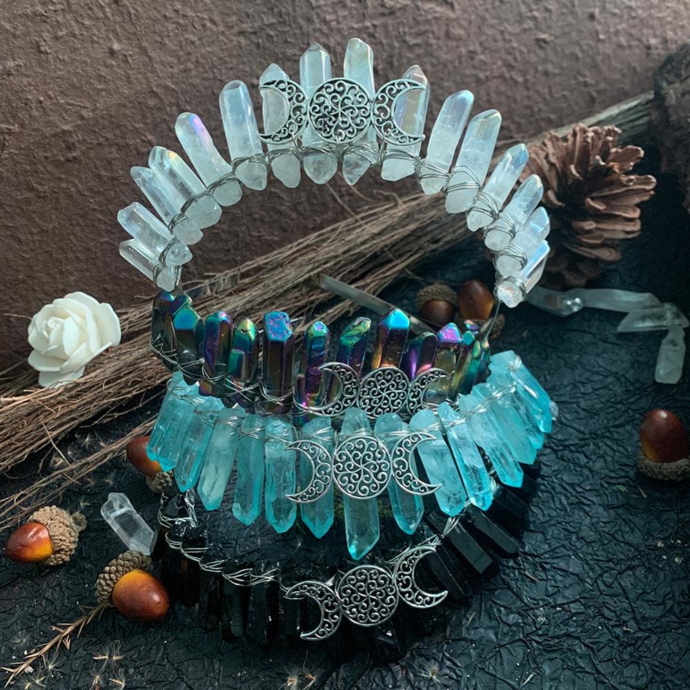 Clear, metallic rainbow, aqua and black crystal crowns stacked on a dark display surface, with silver-tone moon emblems facing forward.