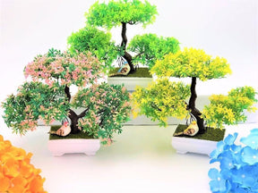 Pink, green and yellow artificial bonsai trees displayed together, each with a dark branching trunk and white footed pot.