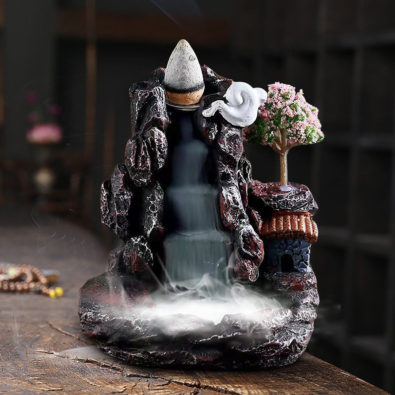 Front view of the village burner with a dense white layer of smoke in the bottom basin. The pink blossom tree and warm roof color contrast with the dark rock-like body.