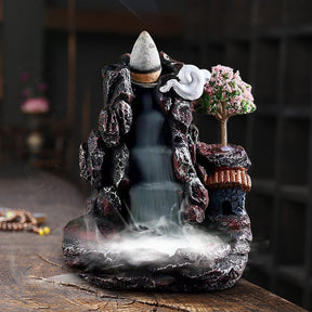 Front view of the village burner with a dense white layer of smoke in the bottom basin. The pink blossom tree and warm roof color contrast with the dark rock-like body.