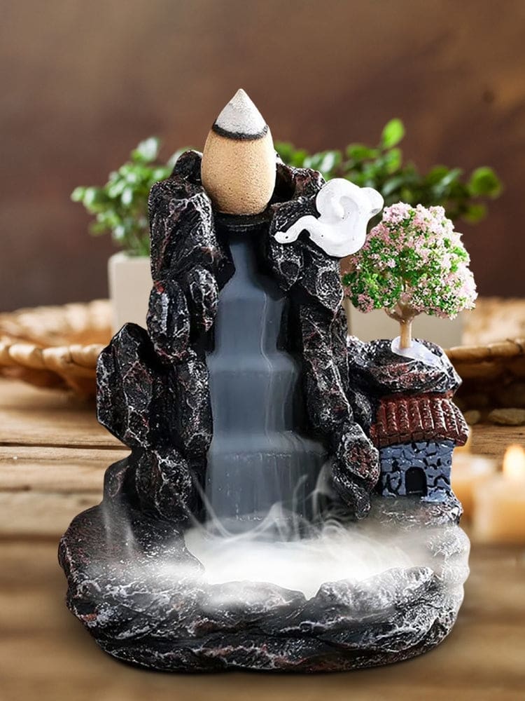 Close front view of the miniature village burner, with white smoke pooling below the rocky stepped channel. A tiny house and pink blossom tree sit along the right side.