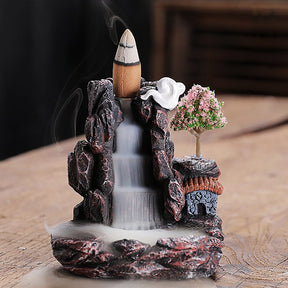 Straight-on view of the burner’s stepped central channel, tiny roofed house and pink tree, with a thin stream of smoke from the cone above.