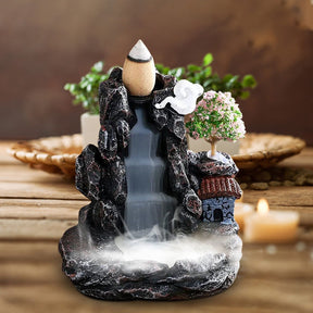Front view of the village backflow burner on a woven mat, with smoke spreading across its lower basin and a blurred plant in the background.