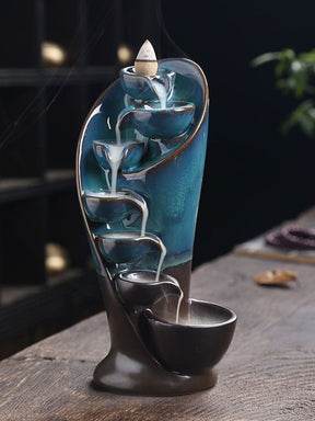 Blue and dark brown waterfall incense burner on a wooden table, with white smoke flowing from the top cone through a descending row of bowls.