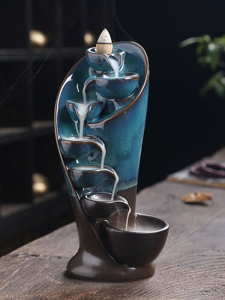 Blue and dark brown waterfall incense burner on a wooden table, with white smoke flowing from the top cone through a descending row of bowls.