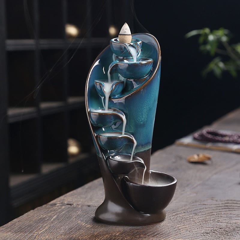 Front view of the blue waterfall burner on wood, showing its curved back and stepped smoke path against a dark interior.