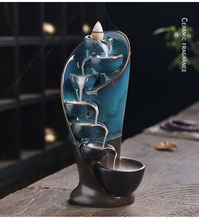 Blue waterfall incense burner shown from the front on a wooden table, with smoke descending through the bowls and a ceramic-fragrance label at the side.