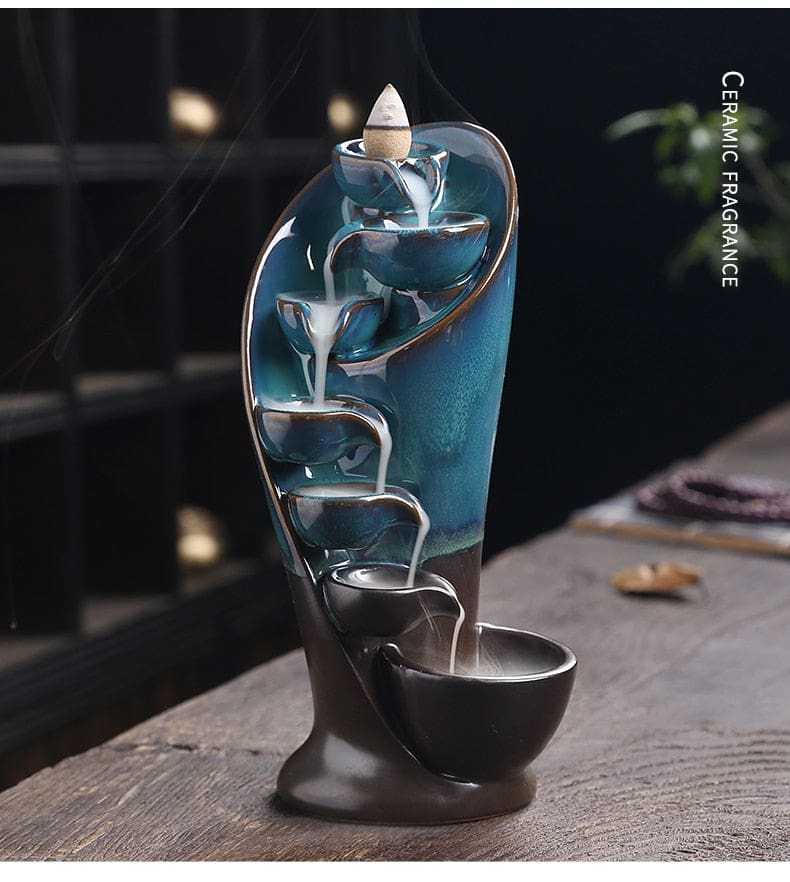 Blue waterfall incense burner shown from the front on a wooden table, with smoke descending through the bowls and a ceramic-fragrance label at the side.