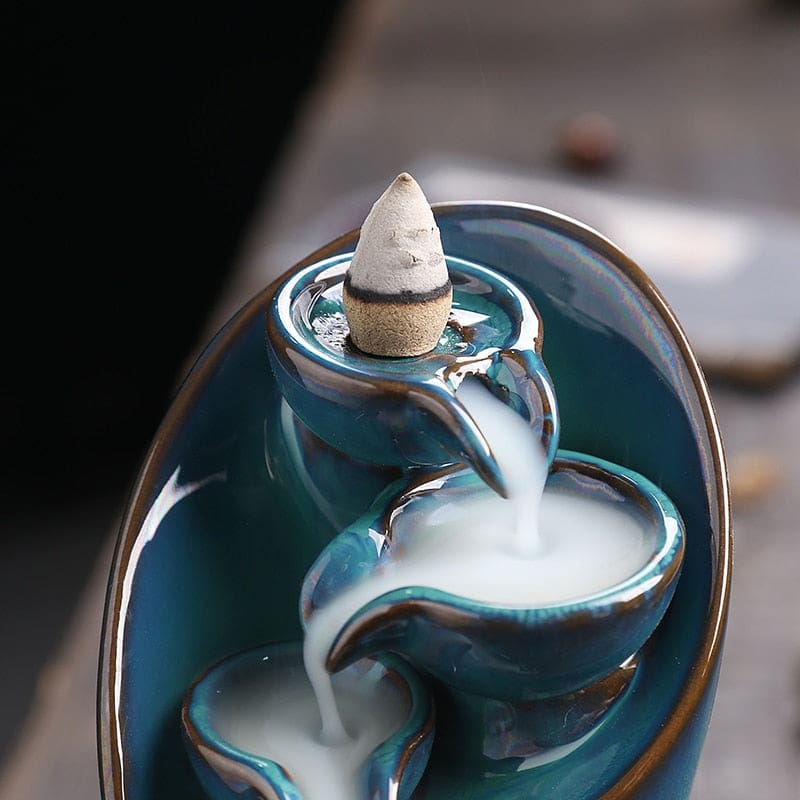 Incense cone seated at the top of the blue backflow burner above a glossy curved bowl filled with smoke.