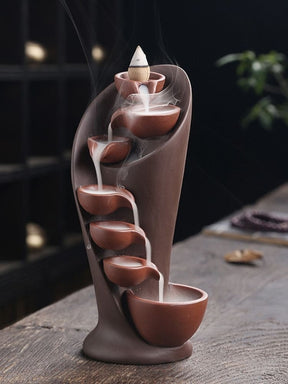 Brown waterfall backflow incense burner with a lit cone at the top, shown as smoke cascades into the lowest bowl.