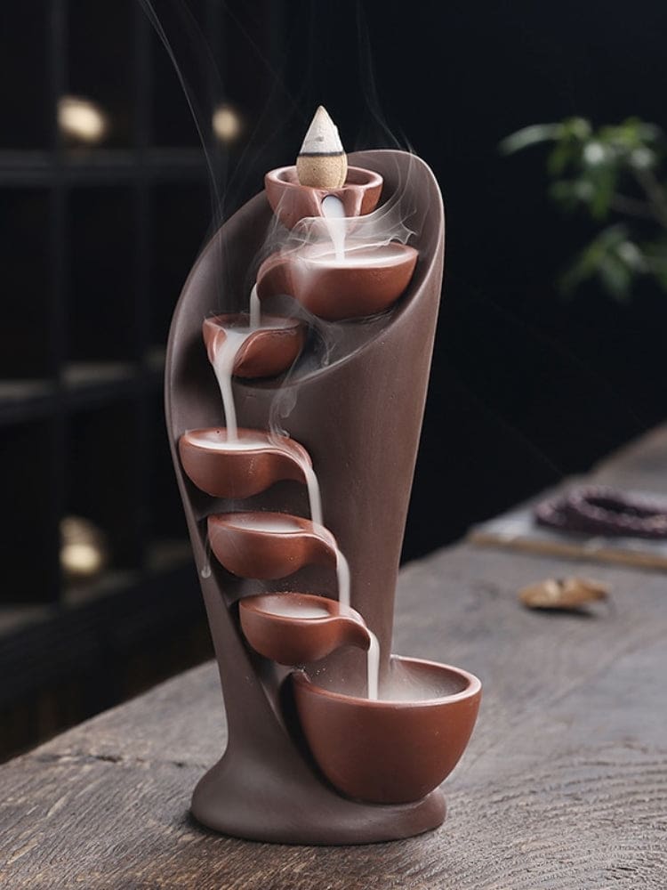 Brown waterfall backflow incense burner with a lit cone at the top, shown as smoke cascades into the lowest bowl.