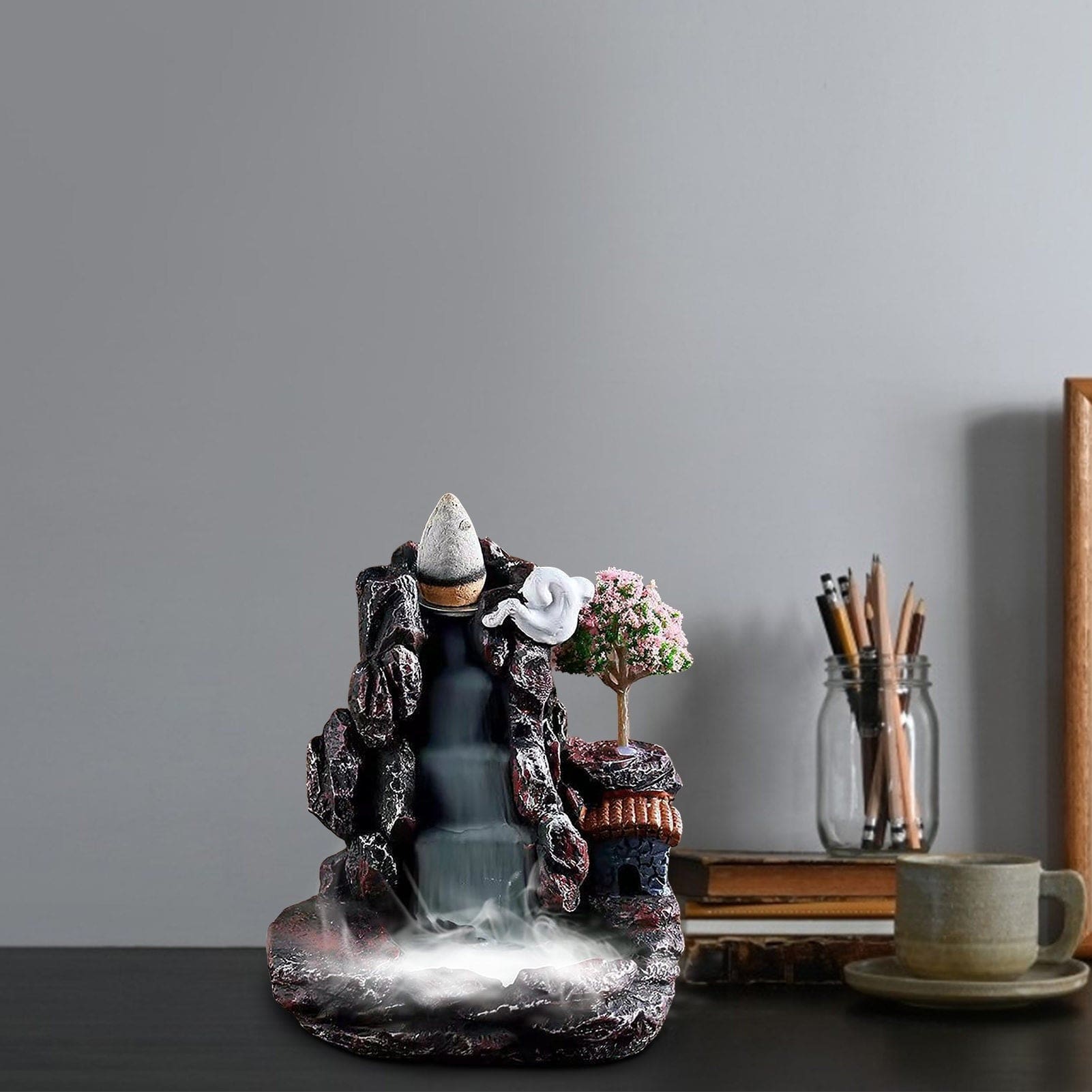 Village waterfall incense burner on a desk with books and a pencil container nearby, its dark rocky outline framing the smoke channel and tiny house.
