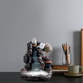 Village waterfall incense burner on a desk with books and a pencil container nearby, its dark rocky outline framing the smoke channel and tiny house.
