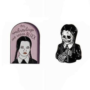 Two braided portrait enamel pins on white: a pink arched We Wear Black design and a black-and-white skeleton figure.